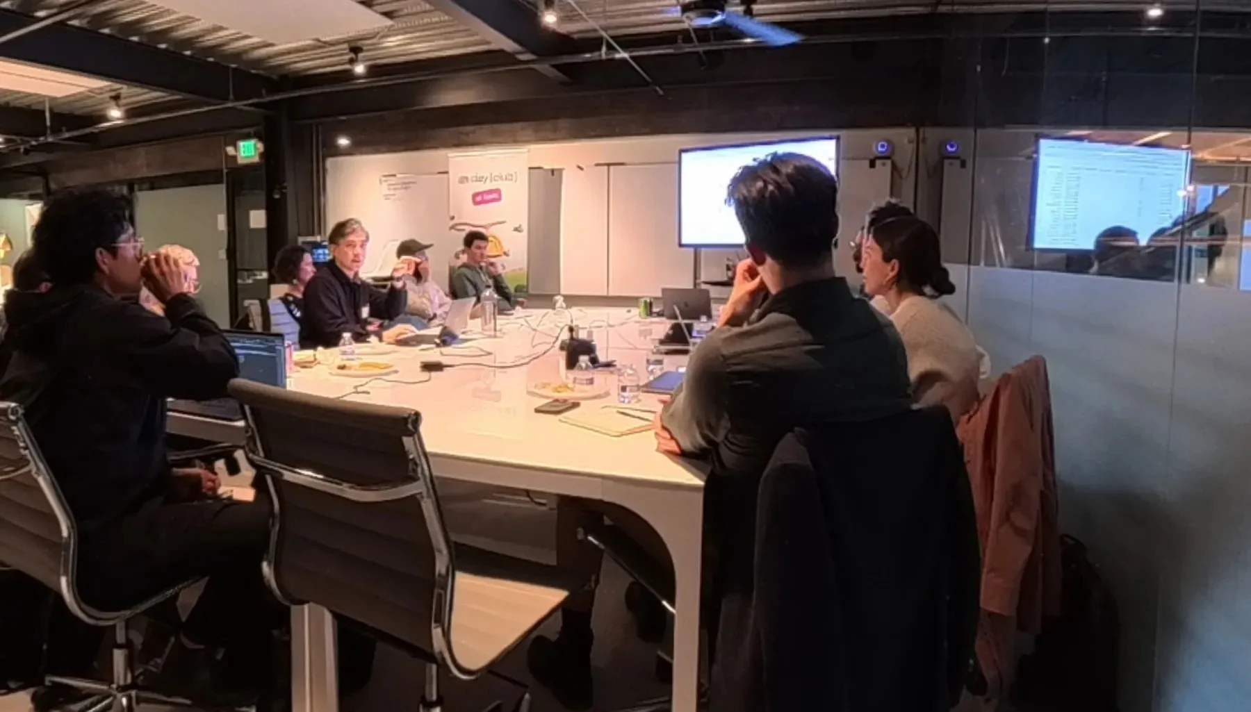 A go-to-market working session around a long table, two screens up, one person mid-sentence and the rest of the room listening