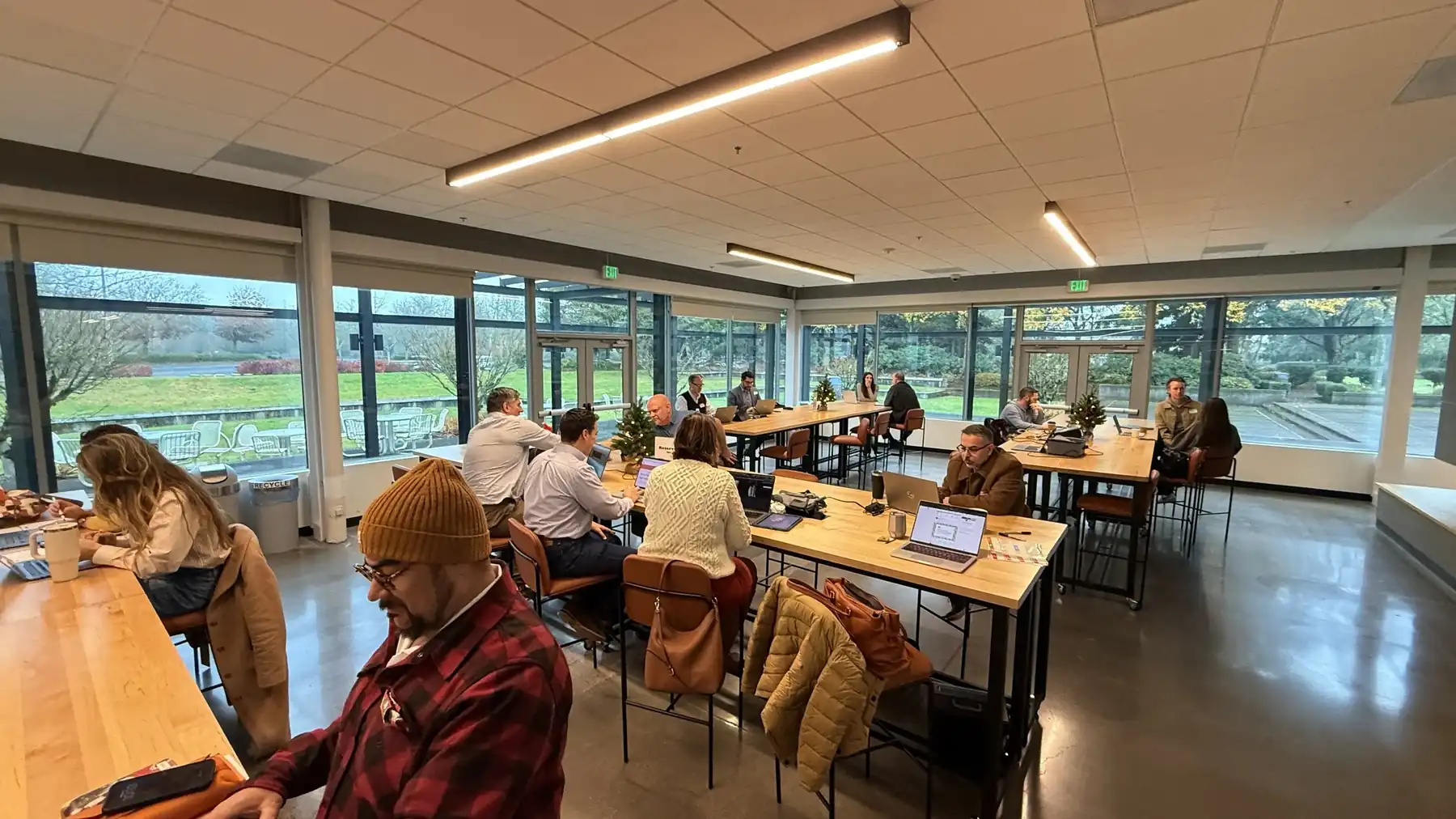 A workshop room of people at long tables with laptops open, floor-to-ceiling windows along one wall looking out to trees