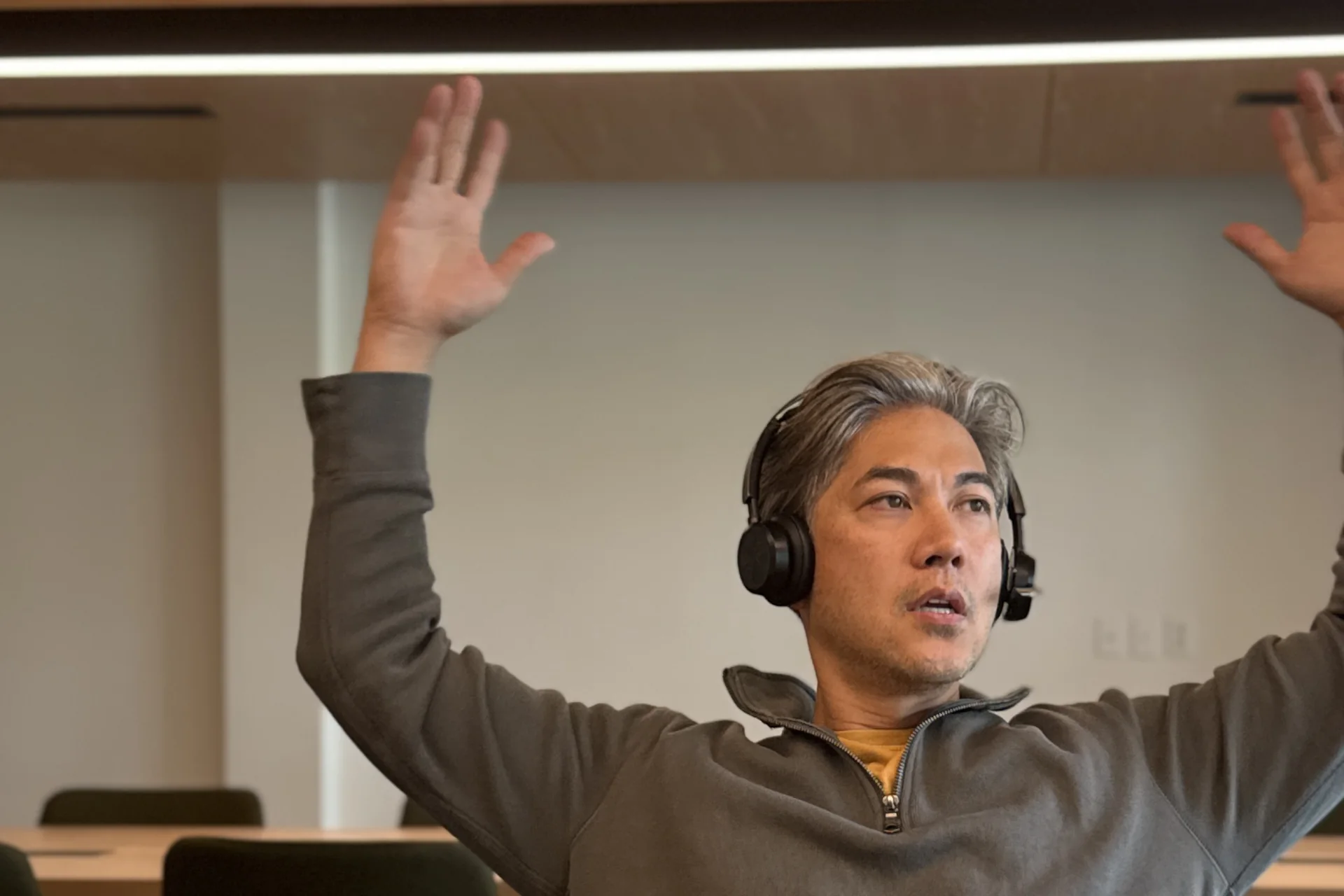Patrick Santiago at a laptop in an empty meeting room, wearing a headset, both arms raised